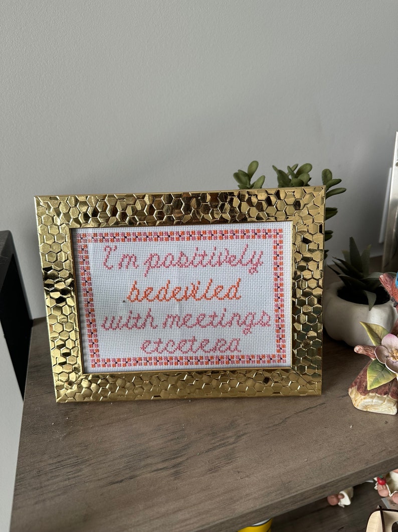May include: A gold-colored frame with a white background and orange and pink text that reads "I'm positively bedeviled with meetings, etcetera."