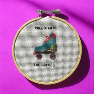May include: Cross-stitch embroidery in a wooden hoop featuring a teal roller skate with red laces and purple wheels. The text "ROLLIN WITH" is above the skate, and "THE HOMIES" is below. The background is a light gray fabric.
