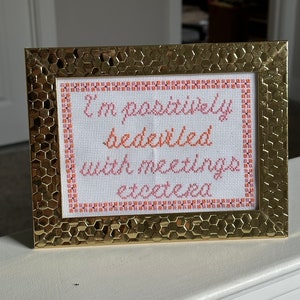 May include: A gold-colored frame with a white background and orange and pink embroidery that reads "I'm positively bedeviled with meetings etcetera."