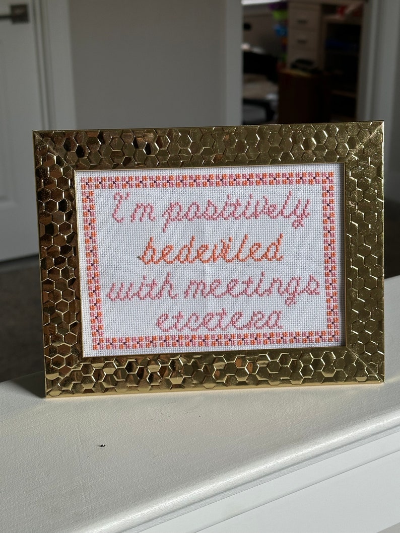 May include: A gold-colored frame with a white background and orange and pink embroidery that reads "I'm positively bedeviled with meetings etcetera."