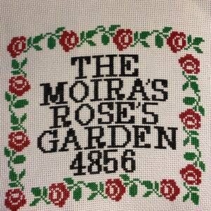 May include: Cross-stitch embroidery with a white background and black text that reads "The Moira's Rose's Garden 4856". The text is surrounded by a border of red roses and green leaves.