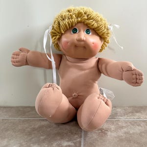 May include: A vintage Cabbage Patch Kid doll with a tan body, blonde yarn hair, and green eyes. The doll has rosy cheeks and is sitting with its arms outstretched. White ribbons are tied in the hair.