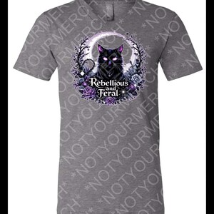 May include: A heather grey short-sleeved t-shirt featuring a graphic of a black cat with glowing purple eyes, surrounded by a circular frame of purple roses and dark foliage, with the text "Rebellious and Feral" printed in white below the cat.
