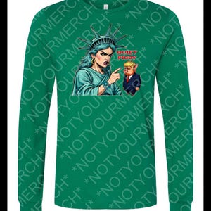 Peut inclure: T-shirt vert &agrave; manches longues avec un dessin de la Statue de la Libert&eacute; pointant vers un cochon en costume, avec le texte "QUIET PIGGY". Le t-shirt porte le texte "NOT YOUR MERCH" r&eacute;p&eacute;t&eacute;.