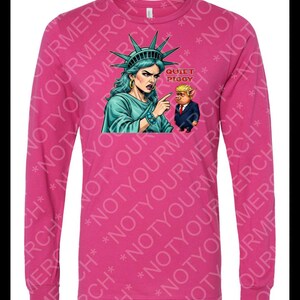 Peut inclure: Un t-shirt rose &agrave; manches longues avec un dessin de la Statue de la Libert&eacute; pointant vers un cochon en costume, avec le texte "QUIET PIGGY". La Statue de la Libert&eacute; est turquoise, et le cochon est en costume.