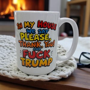 May include: White ceramic mug with a large handle, featuring the text "IN MY HOUSE WE SAY PLEASE, THANK YOU! AND FUCK TRUMP" in colorful, bold lettering. The mug sits on a wooden surface.