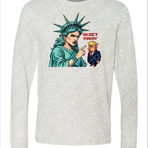 Peut inclure: Un t-shirt gris clair &agrave; manches longues avec un dessin de la Statue de la Libert&eacute; pointant vers un cochon en costume. Le texte "QUIET PIGGY" est au-dessus du cochon. La Statue de la Libert&eacute; est repr&eacute;sent&eacute;e avec la peau et les cheveux bleus.