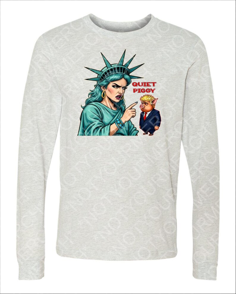 Peut inclure: Un t-shirt gris clair &agrave; manches longues avec un dessin de la Statue de la Libert&eacute; pointant vers un cochon en costume. Le texte "QUIET PIGGY" est au-dessus du cochon. La Statue de la Libert&eacute; est repr&eacute;sent&eacute;e avec la peau et les cheveux bleus.