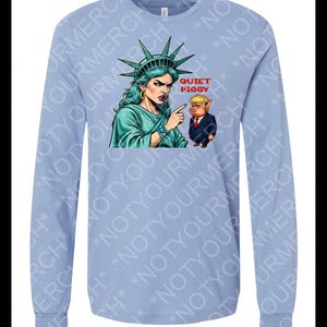 Peut inclure: T-shirt bleu clair &agrave; manches longues avec un dessin de la Statue de la Libert&eacute; pointant vers un cochon en costume. Le texte "QUIET PIGGY" est au-dessus du cochon. Col rond.