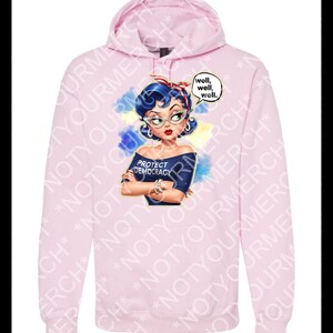 May include: A pale pink hoodie with a graphic of a woman with blue hair, glasses, and a red headband. The graphic includes the text "PROTECT DEMOCRACY" and speech bubbles saying "well, well, well."