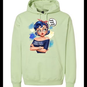 May include: A pale green hoodie with a graphic of a woman with blue hair, glasses, and a red and white headband. The woman is wearing a navy blue off-the-shoulder top that says "PROTECT DEMOCRACY." A speech bubble says "well, well, well."