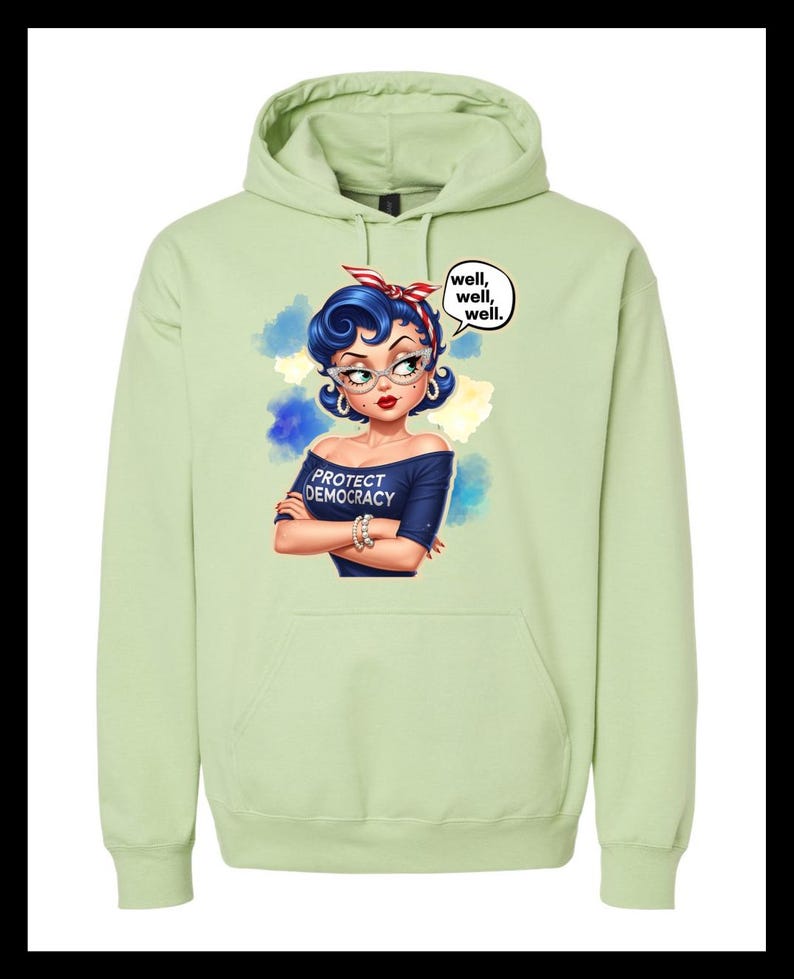 May include: A pale green hoodie with a graphic of a woman with blue hair, glasses, and a red and white headband. The woman is wearing a navy blue off-the-shoulder top that says "PROTECT DEMOCRACY." A speech bubble says "well, well, well."