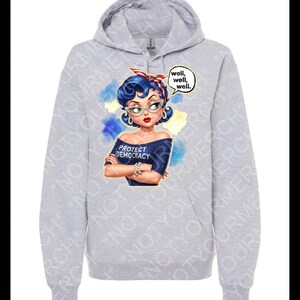 May include: A pale grey hoodie with a cartoon image of a woman with blue hair, glasses, and a red and white headband. The woman wears a blue off-the-shoulder top with the text "PROTECT DEMOCRACY." A speech bubble says "well, well, well."