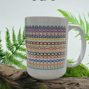 May include: White ceramic mug with a colorful geometric pattern in shades of blue, orange, yellow, red, and purple. The mug has a white handle and the letters "FDT" printed at the top. The mug is displayed on a piece of wood.