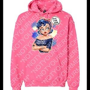 May include: Bright pink hoodie with a graphic of a woman with blue hair, glasses, and a red headband. The woman is wearing a blue shirt that says "PROTECT DEMOCRACY." A speech bubble says "well, well, well."