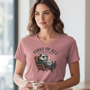 May include: A dusty rose V-neck t-shirt with the text "First of All F*ck Canks". The shirt features a graphic of a badger wearing sunglasses, seated in a chair, and holding a mug with "FDT" printed on it.