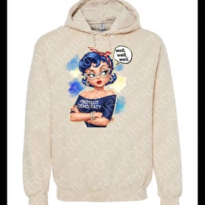 May include: A beige hoodie with a graphic of a woman with blue hair, glasses, and a red headband. The woman is wearing a blue off-the-shoulder top with the text "PROTECT DEMOCRACY." A speech bubble says "well, well, well."
