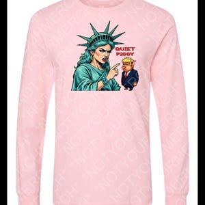 Peut inclure: Un t-shirt rose clair &agrave; manches longues avec un dessin de la Statue de la Libert&eacute; pointant vers un cochon en costume. Le texte "QUIET PIGGY" est au-dessus du cochon. Le t-shirt est &agrave; col rond.