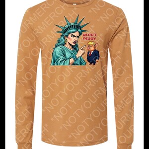 Peut inclure: Un t-shirt &agrave; manches longues de couleur beige avec un motif de la Statue de la Libert&eacute; pointant vers un cochon en costume. Le texte "QUIET PIGGY" est au-dessus du cochon. Le t-shirt porte l'inscription "NOT YOUR MERCH".