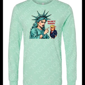 Peut inclure: T-shirt &agrave; manches longues vert menthe avec un dessin de la Statue de la Libert&eacute; pointant vers un cochon en costume. Le texte "QUIET PIGGY" est au-dessus du cochon. Le t-shirt a un col rond et des manches longues.