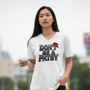 May include: White V-neck t-shirt with the text "DON'T BE A PATSY" in a bold, stylized font. The design includes a red baseball cap graphic. The shirt is worn by a person holding a coffee cup.