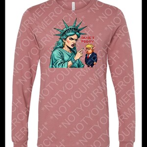 Peut inclure: T-shirt &agrave; manches longues de couleur mauve avec un motif de la Statue de la Libert&eacute; pointant vers un cochon en costume. Le texte "QUIET PIGGY" est au-dessus du cochon. Le t-shirt a un col rond et des manches longues.