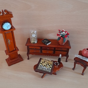 May include: A miniature dollhouse furniture set with a wooden clock tower, a chest of drawers, a chessboard, a small table with a bottle of wine and a glass, and a small stool with a book on top. The set is decorated with a small vase of red flowers.