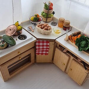 Puede incluir: Un juego de cocina en miniatura para casa de muñecas con armarios de madera, una encimera blanca y varios alimentos en miniatura, incluyendo una olla de sopa, un cuenco de fruta, una cesta de huevos y una tabla de cortar con verduras.