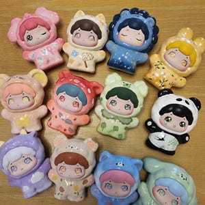 Op de afbeelding: Een verzameling van 12 keramische beeldjes, elk met een schattig cartoonfiguurtje dat een ander dierenkostuum draagt. De beeldjes zijn in verschillende kleuren geschilderd, waaronder roze, blauw, groen, geel en wit. Ze staan allemaal op een houten oppervlak.