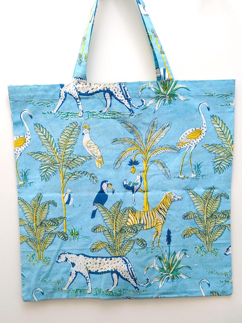 May include: Light blue tote bag with a repeating pattern of jungle animals and plants. The design includes leopards, zebras, toucans, and flamingos. The bag has two matching blue handles.