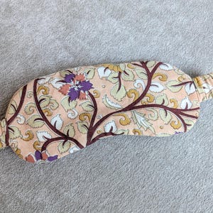 May include: A floral patterned sleep mask in peach, cream, brown, and purple. The mask features an elastic band and is designed to block out light. The fabric has a botanical design with leaves and flowers.