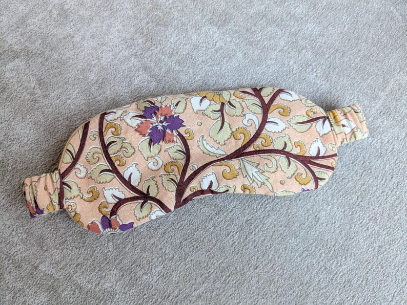 May include: A floral patterned sleep mask in peach, cream, brown, and purple. The mask features an elastic band and is designed to block out light. The fabric has a botanical design with leaves and flowers.