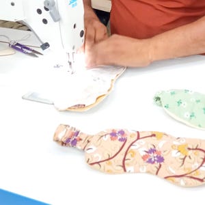May include: A sewing machine in use, stitching a beige fabric with a floral pattern. Several eye masks are visible, one with a floral design, another in green with white flowers. The scene suggests handmade sleep masks.