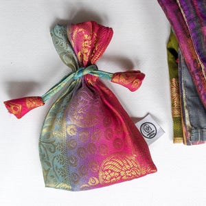 May include: A small drawstring bag in shades of pink, purple, and teal, adorned with gold floral patterns. The bag is tied with a teal cord, and the ends of the cord have pink and gold detailing. A small white tag is attached to the bag.