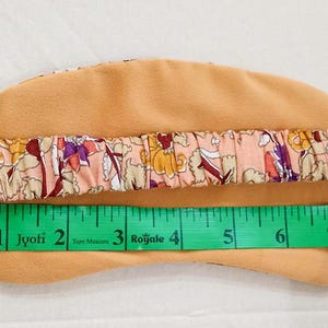 May include: A beige sleep mask with a floral patterned elastic band. The mask is measured at approximately 8 inches in length. The band features a design with pink, purple, and yellow flowers.