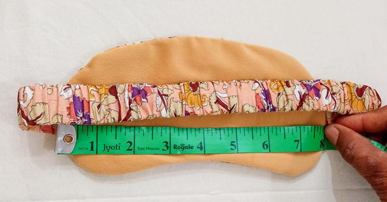 May include: A beige sleep mask with a floral patterned elastic band. The mask is measured at approximately 8 inches in length. The band features a design with pink, purple, and yellow flowers.