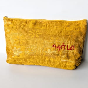 May include: A mustard yellow cosmetic bag with a floral design and a zip. The bag features red embroidered text. The fabric has a textured finish. The bag is a rectangular shape.