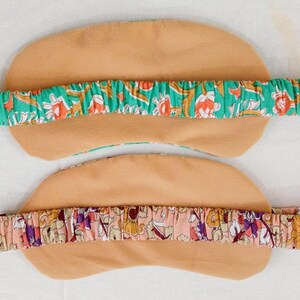 May include: Two eye masks with elastic headbands. The masks are tan, with a floral patterned headband in green, orange, and white. The second mask has a pink headband with a floral pattern in purple, yellow, and red.