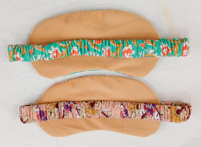 May include: Two eye masks with elastic headbands. The masks are tan, with a floral patterned headband in green, orange, and white. The second mask has a pink headband with a floral pattern in purple, yellow, and red.