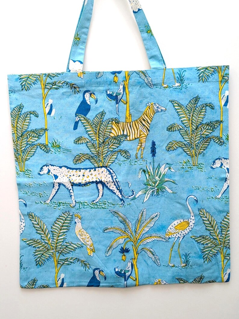 May include: A blue tote bag featuring a jungle scene with zebras, leopards, toucans, and palm trees. The bag has blue handles and a repeating pattern of the jungle animals and foliage. The background is a light blue color.
