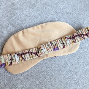 May include: A beige sleep mask with a floral patterned elastic band. The mask is made of a soft material and is designed to block out light. The elastic band features a pattern of brown, purple, and green floral elements.