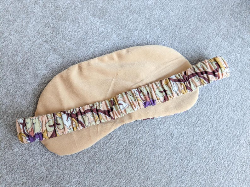 May include: A beige sleep mask with a floral patterned elastic band. The mask is made of a soft material and is designed to block out light. The elastic band features a pattern of brown, purple, and green floral elements.