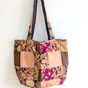 May include: A patchwork tote bag featuring a mix of patterned fabric squares in brown, tan, and purple hues. The bag has long straps with a black interior, set against a white backdrop. Designed for carrying items, it's a stylish accessory.