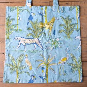 May include: Light blue tote bag with a repeating pattern of jungle animals and plants. The design includes a zebra, leopard, toucans, and palm trees in yellow, white, and blue. The bag has two handles.