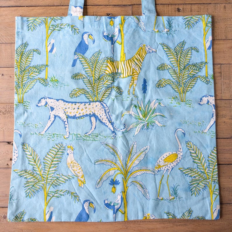 May include: Light blue tote bag with a repeating pattern of jungle animals and plants. The design includes a zebra, leopard, toucans, and palm trees in yellow, white, and blue. The bag has two handles.