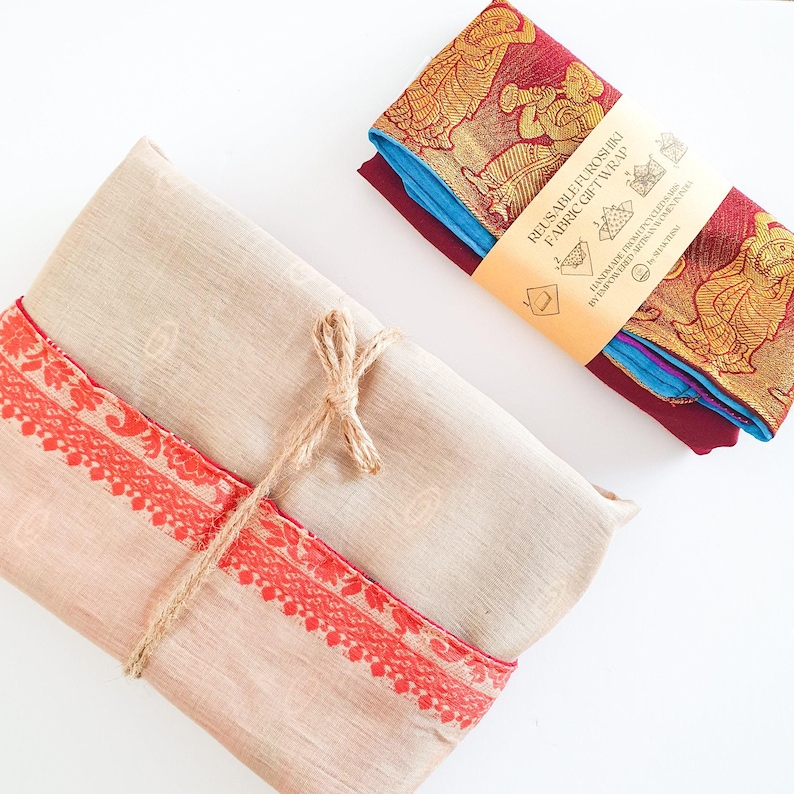 May include: A beige fabric gift wrap with a red and gold border is tied with twine. The wrap is folded and ready to be used. A separate label with the text "Reusable Furoshiki Fabric Gift Wrap" is included.