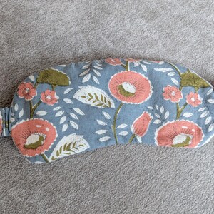 May include: A floral sleep mask with a light blue background. The mask features a repeating pattern of large coral-colored flowers with white and green leaves. The elastic band is made of the same floral fabric.