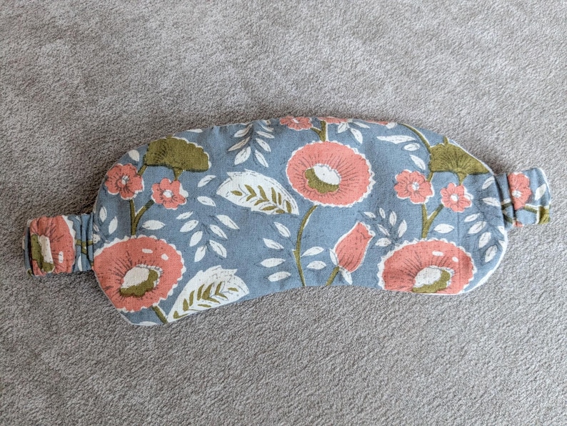 May include: A floral sleep mask with a light blue background. The mask features a repeating pattern of large coral-colored flowers with white and green leaves. The elastic band is made of the same floral fabric.