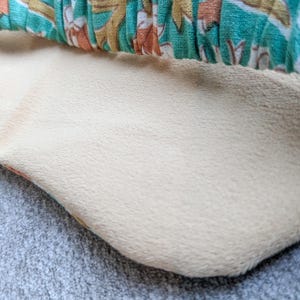 May include: Close-up of a cream-colored, plush fabric with a soft texture. The fabric is bordered by a patterned teal, orange, and white fabric. The image is set against a gray, textured surface.
