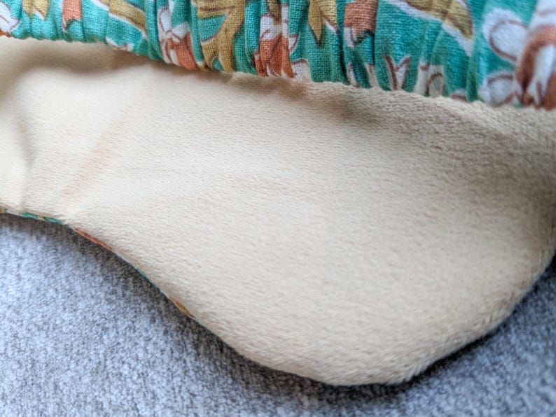 May include: Close-up of a cream-colored, plush fabric with a soft texture. The fabric is bordered by a patterned teal, orange, and white fabric. The image is set against a gray, textured surface.